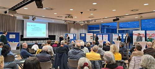 In a large hall, two professors present the results of the online survey to a large audience using a projector and posters.