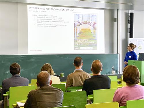 Ein Vorlesungssaal mit einer Referentin, die zu Fachpublikum spricht. Die Sitzbänke und Tische sind aus Holz und in Variationen der Farbe Grün gefärbt. Es ist eine projizierte Präsentation von einem Beamer zu sehen, die das integrierte Klimaschutzkonzept der Hochschule darstellt. Die Referentin ist in der rechten Bildseite zu finden, zur Beamerfläche gedreht und trägt einen knallblauen Wollpulli. Der Raum ist hell und umringt von Sichtbetonwänden. Die grüne Kreide-Schreibtafel ist herunter geschoben worden. Im Vordergrund befindet sich das Publikum bestehend aus 2 Frauen und 4 Männern, die allesamt von hinten abgebildet sind.