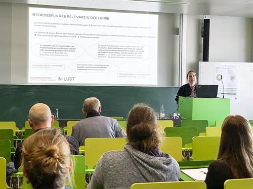 Ein Vorlesungssaal mit einer Referentin, die zu Fachpublikum spricht. Die Sitzbänke und Tische sind aus Holz und in Variationen der Farbe Grün gefärbt. Es ist eine projizierte Präsentation von einem Beamer zu sehen, die die interdisziplinäre Lehre der Hochschule darstellt. Die Referentin ist in der rechten Bildseite zu finden und trägt einen weite Jacke. Der Raum ist hell und umringt von Sichtbetonwänden. Die grüne Kreide-Schreibtafel ist herunter geschoben worden. Im Vordergrund befindet sich das Publikum bestehend aus 2 Frauen und 3 Männern, die allesamt von hinten abgebildet sind.