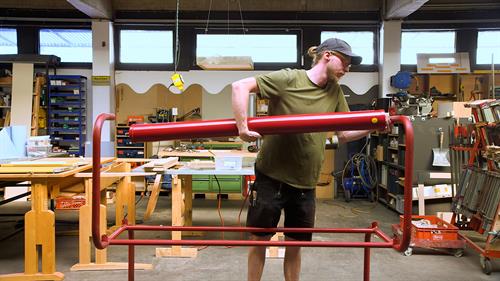 Eine männliche Person mit Kappy und längeren Haaren zum Zopf werkelt an einem knallroten Metallgestänge. Er trägt ein khakifarbenes T-Shirt und eine kurze schwarze Hose mit einiges an Werkzeug in den Taschen. im Hintergrund ist die Werkstatt abgebildet.