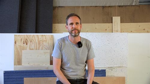 Abgebildet ist ein Mann mittleren Alters, der mittig im Bild sitzt. Er trägt kurze braun-graue Haare und einen Bart. Außerdem trägt er ein einfaches, kurzärmeliges T-Shirt und eine schwarze Jeans. Im Hintergrund stehen aufrecht unterschiedliche Formate von verschiedenen Holzplatten.