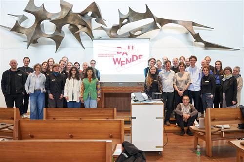 Eine große Gruppe von diversen Menschen stehen zur Kamera gedreht in drei Reihen. In der Mitte ist ein Projektor, der die Überschrift "Hochschulnetzwerk – Gemeinsam für die Bauwende" auf eine Leinwand wirft. Darüber befindet sich eine stachelige metallische Skulptur, die die komplette Wand ziert. Der Boden des Raumen ist aus Holz und im Vordergrund stehen Holzbänke, wie in einer Kirche.