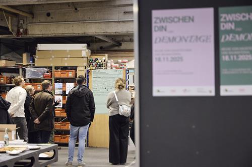 Auf der linken Seite des Bildes ist der Einblick einer Werkstatt mit einem Halbkreis aus Menschen, die alle auf ein hellgrünes Plakat blicken. Die rechte Bildhälfte ist dunkelbraun. Darauf befinden sich zwei Plakate. Das eine in rosa mit dunkelgrüner Schrift, das andere in dunkelgrün mit rosa Schrift.