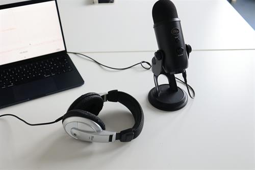 Abgebildet ist ein Tisch mit einem Laptop links in der Ecke, rechts davon ist ein kleines Standmikrofon zur Aufnahme und On-Ear-Kopfhörer mit schwarzen dicken Polstern. Das Foto ist mit leichter Aufsicht fotografiert.