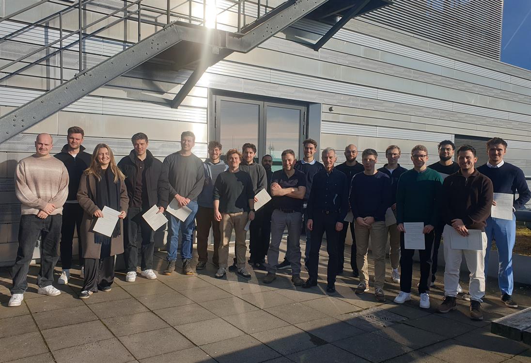 Eine diverse Gruppe an Studierenden und Lehrenden stehen bei Sonnenschein auf einer Terrasse der Hochschule Düsseldorf mit dem Gesicht zur Kamera.