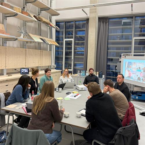 Eine Gruppe von 5 Frauen und 4 Männern sitzt um einen großen Tisch im Kreis und sind in eine Diskussion vertieft. Im Hintergrund steht ein sehr hohes Holzregal mit jeglichen Materialien rund um das Thema Nachhaltigkeit/zirkuläres Bauen. Im Hintergrund befinden sich bodentiefen Fenster mit schweren Vorhängen und ein Beamer wirft die Online-Teilnehmer*innen auf ein Whiteboard.