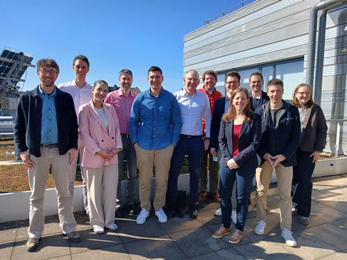 Eine Gruppe von 12 diverser Menschen stehen auf einer Art Terrasse draußen und sind mit Körper und Gesicht zur Kamera gedreht. Es ist ein sonniger Tag. Die Personen auf dem Bild sind leger bis chic gekleidet.