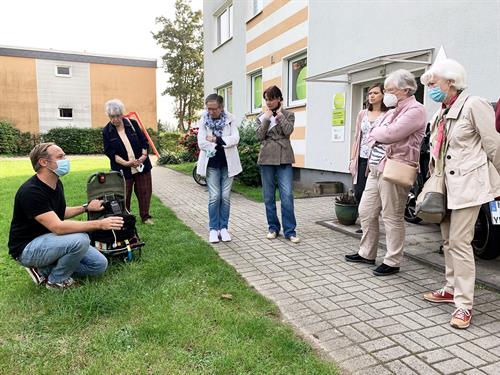 Auf dem Bild zu sehen ist eine kleine Gruppe an Menschen, die vor einem Haus, dem "Quartierspunkt", in Mülheim Heißen-Süd stehen. Vor einem dieser Gruppe steht ein "Rucksack", welcher zur Messung von Schadstoffen in der Luft ist
