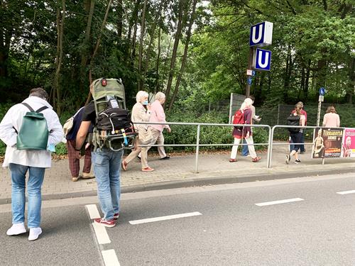 Auf dem Bild zu sehen ist eine kleine Gruppe an Menschen, die eine Straße in Mülheim Heißen-Süd überqueren um so zur U-Bahn Haltestelle "Eichbaum" zu gelangen. Im Vordergrund ist eine Person mit "Rucksack" zu sehen, welcher zur Messung von Schadstoffen in der Luft ist.