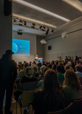 Blick in einen Vortragsraum bei der Nacht der Wissenschaft Düsseldorf. Der Fotograf steht hinten im Raum und hat über die Köpfe der Besucher hinweg die Bühne in den Fokus genommen.