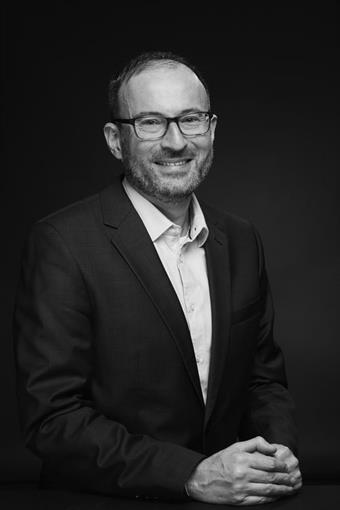 Das Bild zeigt einen lächelnden, leicht schrägsitzenden Herren mit dunkler Brille, kurzen dunklen Haaren und kurzem 3-Tages-Bart. Er trägt ein Sakko und ein weißes Hemd.