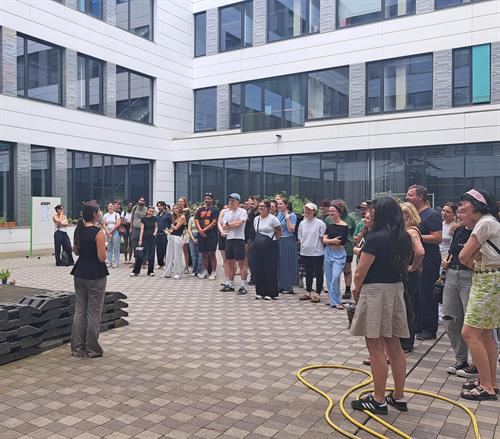 Eine weiblich gelesene Person trägt innerhalb eines Innenhofs für eine diverse Gruppe Studierende vor.