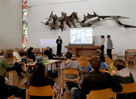 Eine vortragende Studentin steht in einer Kirche vor einer Beamer-Präsentation. Rechts von ihr steht eine weitere Studentin und ein Student. Beide mit dem Körper zu der Vortragenden gedreht.
Im Vordergrund sind viele Holzstühle platziert, die zur kleinen Empore gerichtet sind, auf der die drei Studierenden stehen. Auf diesen Stühlen sitzen Personen verschiedenen Alters.
Der Raum ist sehr hoch und weiß verputzt. Der Boden ein Holzboden und links von der Vortragenden befindet sich ein sehr hohes schmales buntes Kirchenfenster mit bunten, großen Mosaikmustern, welches weiches Licht in den Raum lässt.
Auf der hinteren Wand hängt ein sehr großes dekoratives Objekt aus Metall an der Wand.