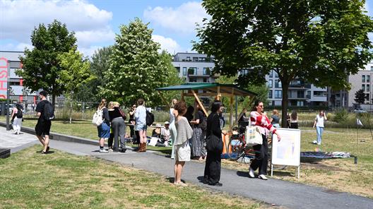 Das Bild zeigt viele Studierende, auf einer Wiese sitzend und plaudernd, umringt von mobilen Möbeln, die an einem Fahrrad angebracht sind und eine Außenküche bilden. Im Hintergrund sind kleinere Bäume und die nahe gelegene Wohnsiedlung mit weißen modernen Wohnkuben zu sehen.