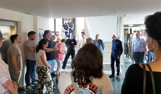 In einem niedrigen Vorraum mit anschließendem Treppenhaus stehen im Kreis Menschen, die dem Vortragenden zuhören und lächeln. Die Gruppe ist divers. Der Raum ist mit abgehangen mit weißen Akustikplatten und der Boden besteht im Durchgangsraum aus grün-marmoriertem Stein. Die Wände des Treppenhaus aus Waschbeton und die Treppen aus einem grauen Naturstein. Das Treppenhaus hat ebenso Fenster, die viel Tageslicht hineinlassen. Ebenso ist der Vorraum lichtdurchflutet.