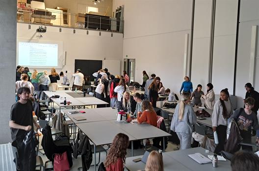 Eine große Menge an Studierenden sitzen an Gruppentischen ausgerichtet zum Professor. Der Vorlesungsraum ist sehr langgezogen und sehr hoch. Die sichtbaren Wände bestehen aus Sichtbeton.