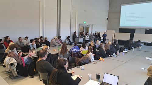 Eine große Menge an Studierenden sitzen an Gruppentischen ausgerichtet zum Professor. Der Vorlesungsraum ist sehr langgezogen und sehr hoch. Die sichtbaren Wände bestehen aus Sichtbeton.