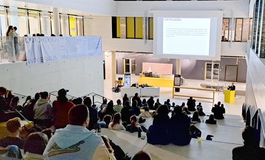 Das große Atrium des Gebäudes 6 der Hochschule Düsseldorf mit vielen Studierenden auf einer breiten Treppe. Vorn steht eine Referentin, die einen Vortrag hält.