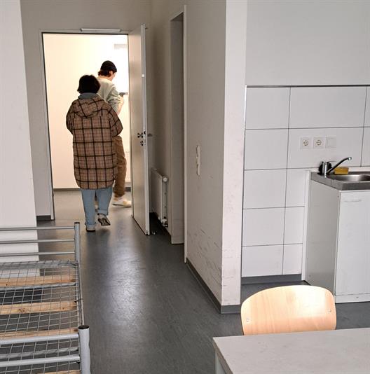 Abgebildet ist eine Ein-Raum-Wohnung. Im linken Bildrand steht ein niedriges Bettgestell in grau ohne Matratze. Im rechten unteren Bild ist ein einfacher grauer Tisch ohne Dekorationen mit dazugehörigem Stuhl aus Holz zu erkennen. In einer kleinen Nische ist eine kleine weiße Küchenzeile mit weißen halbhohem Fliesenspiegel und einem Metallwaschbecken.
Die Wände und Türen sind weiß gestrichen. Der Boden ist aus grauem Linoleum.
In der Eingangstür sind zwei Personen von hinten zu erkennen. Eine von den beiden Personen ist weiblich gelesen mit langem  Wollmantel und eine männlich große gelesene Person, welche eine Mütze trägt.
